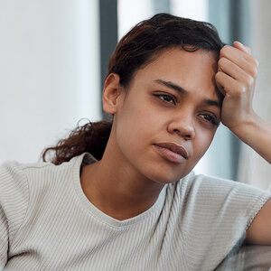 A woman with a worried look sits on a couch with her hand to her forehead.