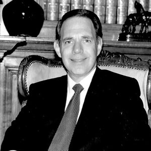 A man in a dark suit sitting in a highbacked chair – an image of Barry Tarlow. A man in a dark suit sitting in a highbacked chair – an image of Barry Tarlow.