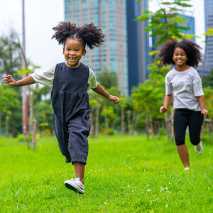 Two young girls run across a grass field in a park with newly planted trees and tall buildings in the background.