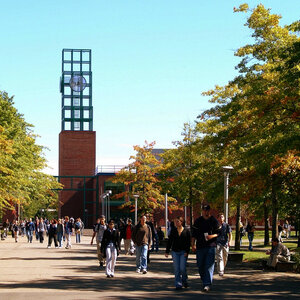 Students walk along a treelined path on a college campus with a modern clocktower in the background.