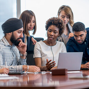 A diverse group of men and women review information on a laptop at a conference table.