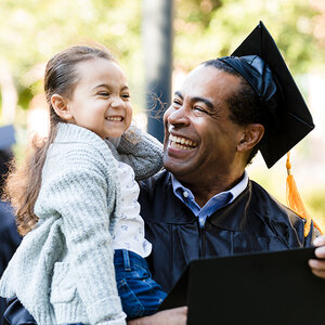 A smiling father wears a cap and gown at his graduation ceremony, while holding his daughter.