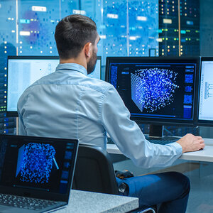 A male data scientist sits in front of a computer screen displaying a three-dimensional representation of data. A male data scientist sits in front of a computer screen displaying a three-dimensional representation of data.
