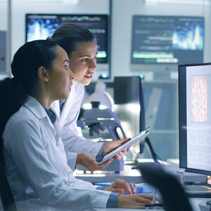 Two women scientists in lab coats looking at a computer screen.
