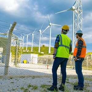 Workers in hard hats and reflective vests stand outside a fenced-in power plant near wind turbines.