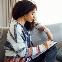 A female African American teenager sits on couch writing a list. A female African American teenager sits on couch writing a list.
