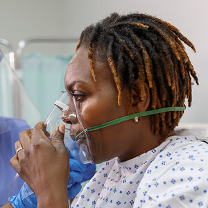 A Black woman in a hospital gown is wearing an oxygen mask to help her breathe.