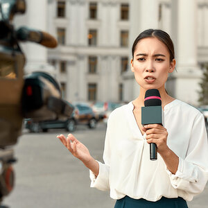 A female TV journalist stands outside a building giving a report before a television camera A female TV journalist stands outside a building giving a report before a television camera