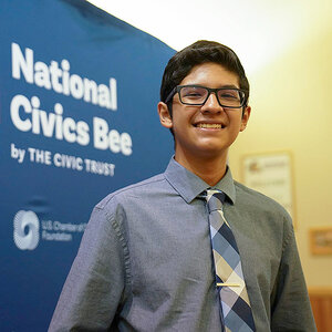 A middle-school aged boy wearing a tie and glasses smiles in front of a sign that reads: National Civics Bee. A middle-school aged boy wearing a tie and glasses smiles in front of a sign that reads: National Civics Bee.