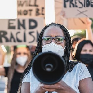 Black woman in glasses holding a megaphone at a protest with "end racism" sign behind her.