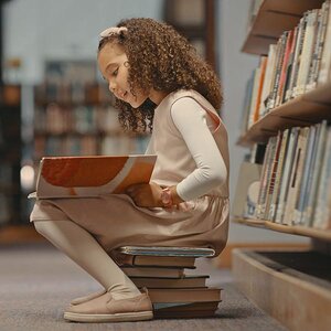 A child sitting on a stack of books reading in a library.