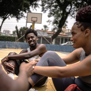 A young African American man and woman sit on a park basketball court smiling and talking.