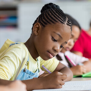 A young African American girl in a classroom writes in her notebook.
