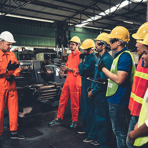 An instructor and six trainees, including a woman of color, in hard hats on a factory floor.
