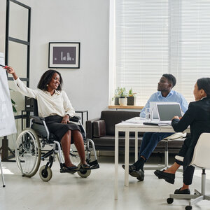 A Black woman in a wheelchair points to a flipchart on an easel in front of work colleagues sitting at a table.