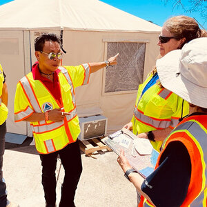 A relief worker wearing a yellow vest stands in front of an emergency shelter directing other relief workers.