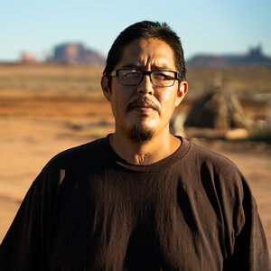 A man in a brown shirt and glasses with a desert and mountains in the background.