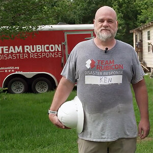 A middle-aged male volunteer is holding a white hardhat and wearing a t-shirt that reads: Team Rubicon Disaster Relief.