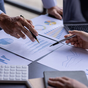 A closeup of people’s hands pointing to charts and graphs on an office desk. A closeup of people’s hands pointing to charts and graphs on an office desk.