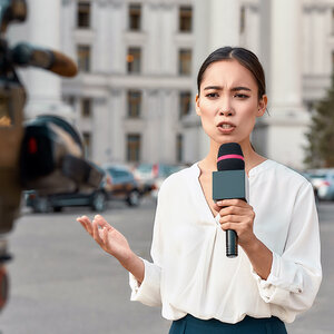 A female reporter outside a building speaks into a microphone in front of a video camera.