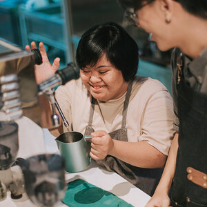 A young woman with developmental disabilities is trained by a barista to operate an espresso machine.