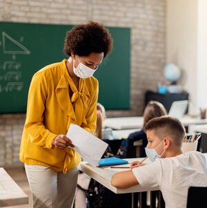 A math teacher shows an elementary school student a worksheet; a geometry problem is on the blackboard.