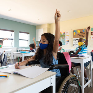 A girl in a wheelchair and wearing a mask raises her hand in a classroom with other students. A girl in a wheelchair and wearing a mask raises her hand in a classroom with other students.