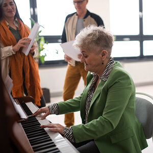 An older woman in a green jacket plays the piano.