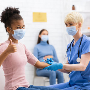 A young woman with a mask receives a vaccination from a nurse, also wearing a mask.