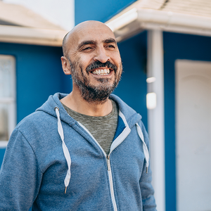 A middle-aged man with a beard wearing a light-blue hoodie is standing in front of a small blue house.