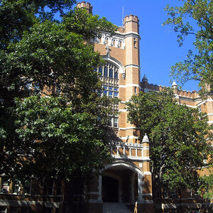 The façade of brick and stone college building surrounded by trees under a blue sky.