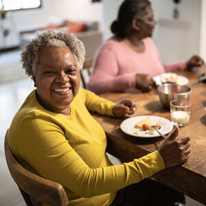 An older African American woman in a yellow sweater is eating a meal at a kitchen table. An older African American woman in a yellow sweater is eating a meal at a kitchen table.