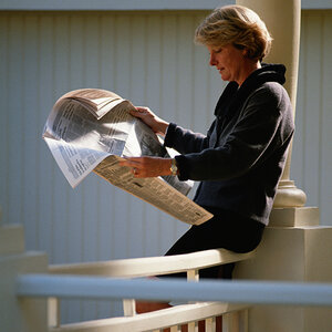 A woman sits on her front railing reading a newspaper.