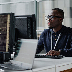 A Black male sits at a computer in a tech office setting.