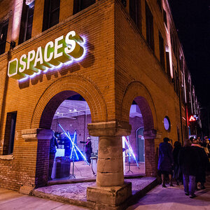 The arched exterior of a brick building at night with a bright sign above that reads: SPACES. The arched exterior of a brick building at night with a bright sign above that reads: SPACES.