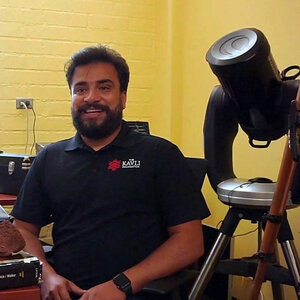 A man with a beard sits at a desk next to a telescope in an office with a yellow wall – a photo of Abraham Amiri.