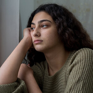 A sad teenage girl with long brown hair rests her head in her hand while staring out a window. A sad teenage girl with long brown hair rests her head in her hand while staring out a window.