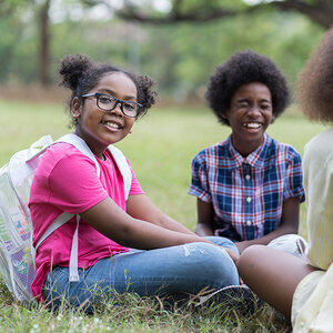Two young African American girls sit in the grass smiling.