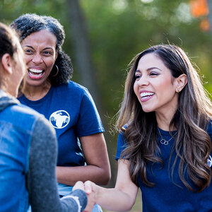 Three female volunteers greet each other at an outdoor event.