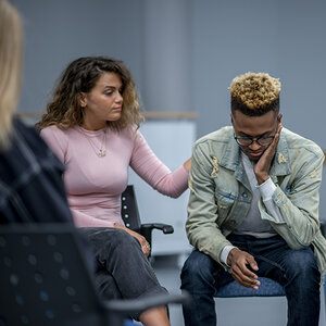 A group session with a Black youth looking down, a woman with a hand on his shoulder.