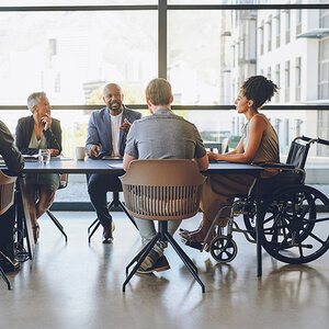 A diverse group of workers sit at a table in an office setting. A diverse group of workers sit at a table in an office setting.