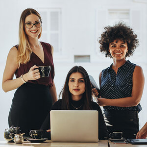 Three women, one seated behind a laptop, pose for a photo at a business meeting.
