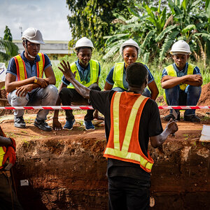 An archeologist and workers review an earthen excavation with palm trees in the background…. An archeologist and workers review an earthen excavation with palm trees in the background….