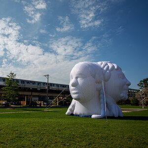 A giant white inflatable sculpture of a woman’s head is tethered to a green lawn with an elevated subway running in the background. A giant white inflatable sculpture of a woman’s head is tethered to a green lawn with an elevated subway running in the background.