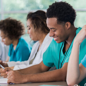 An African American man dressed in hospital scrubs is taking notes in a classroom. An African American man dressed in hospital scrubs is taking notes in a classroom.