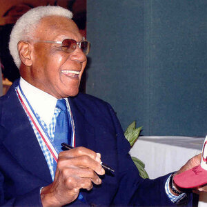 An older African American man with gray hair dressed in a suit an sunglasses signs an autograph—a photo of Buck O’Neil. An older African American man with gray hair dressed in a suit an sunglasses signs an autograph—a photo of Buck O’Neil.