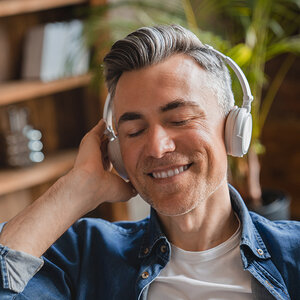 A man with graying hair, eyes closed and smiling, listens to music on his headphones.