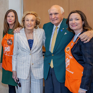 An older husband and wife pose with two women wearing orange aprons—A photo of Elaine and Ken Langone. An older husband and wife pose with two women wearing orange aprons—A photo of Elaine and Ken Langone.