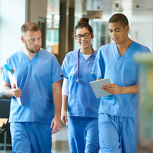 Three medical students in blue scrubs walk down a hallway as they review a chart.
