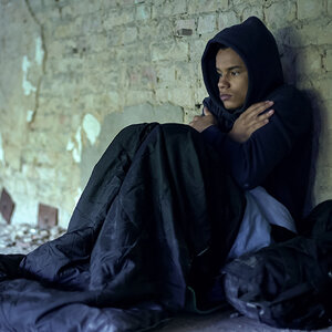A teen in a hoodie sits against a dirty brick wall huddling for warmth.
